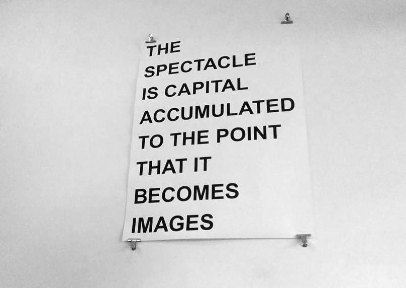 Poster clipped to a white wall: THE SPECTACLE IS CAPITAL ACCUMULATED TO THE POINT THAT IT BECOMES IMAGES